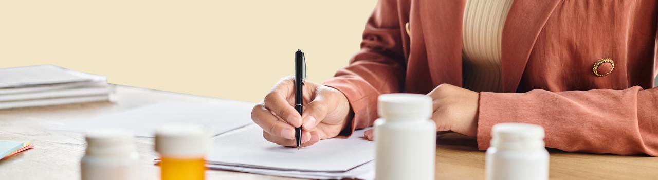 An image showing medicine on the table and a person writing on paper with a black pen.