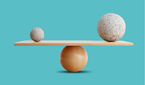 A balancing board teeters on a wooden sphere, with a small stone ball on one end and a larger stone ball on the other. The background is a plain light blue.